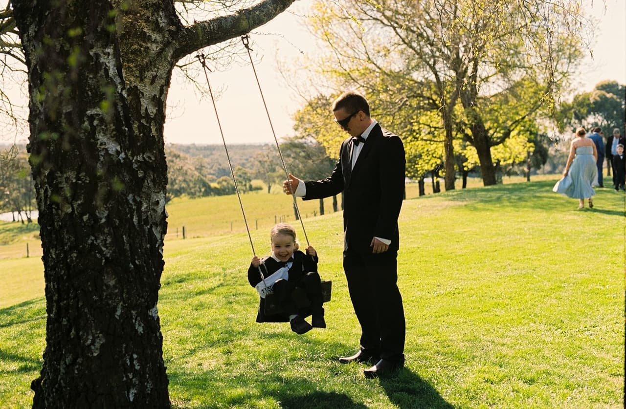 A guest pushing a little boy on a rope swing hanging from a big old tree on the lawn, on 35mm film
