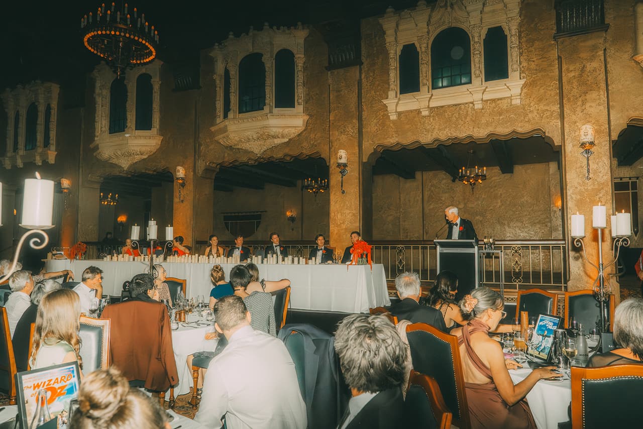 Guests at dinner below the carved balconies and arched windows along the ballroom wall, a speech under way at the bridal table