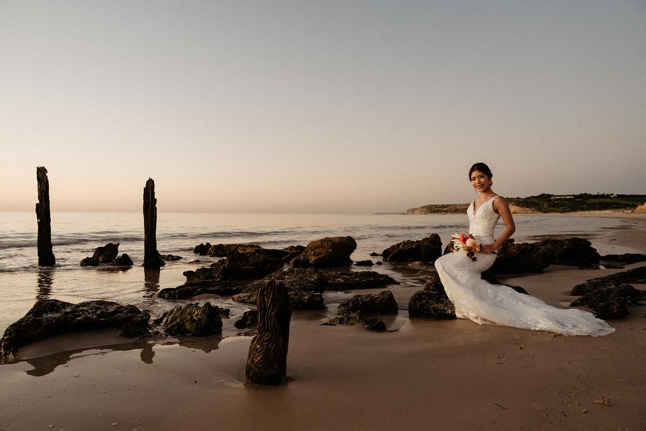 Sayuri sitting on the dark rocks among shallow pools near the jetty posts, the coastline behind her