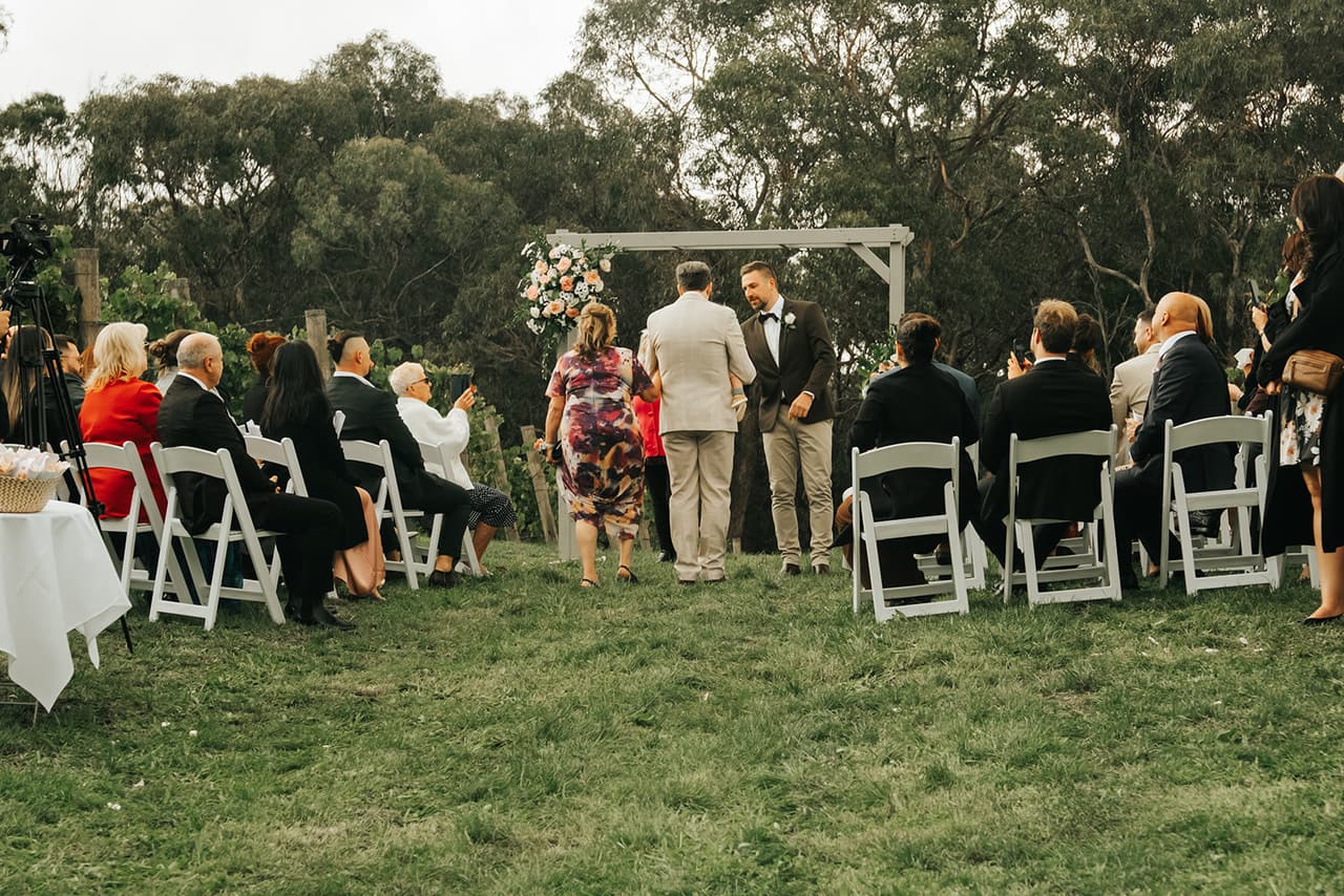 The ceremony seen from behind the seated guests, Max waiting under the flower-trimmed arbour