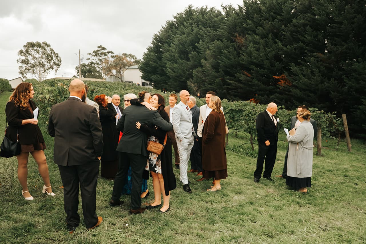 Guests gathering on the grass beside the vines before the ceremony, a cypress hedge and gum trees behind