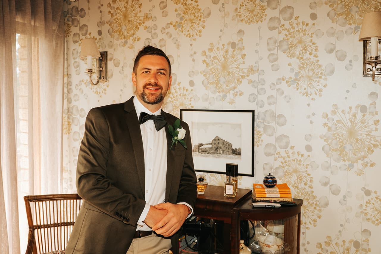 Max sitting in a room with gold floral wallpaper, a wall lamp and a framed drawing of an old barn