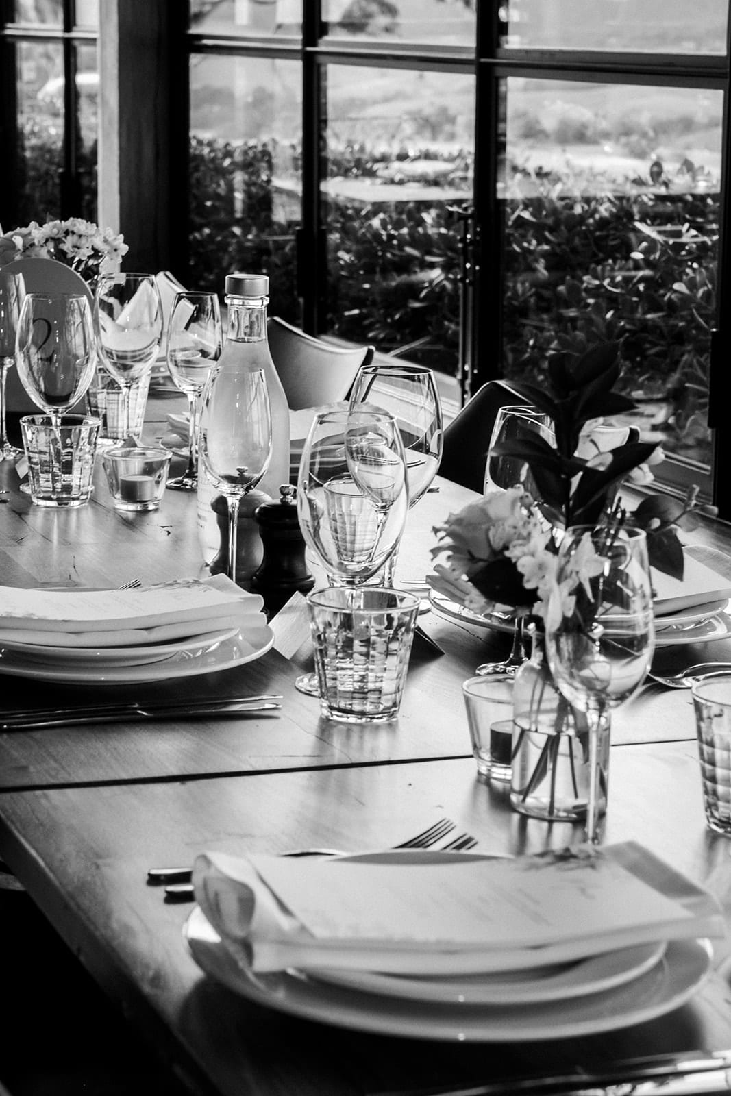 Black and white photo of a place setting with folded napkin, glasses and a small vase of flowers by the window