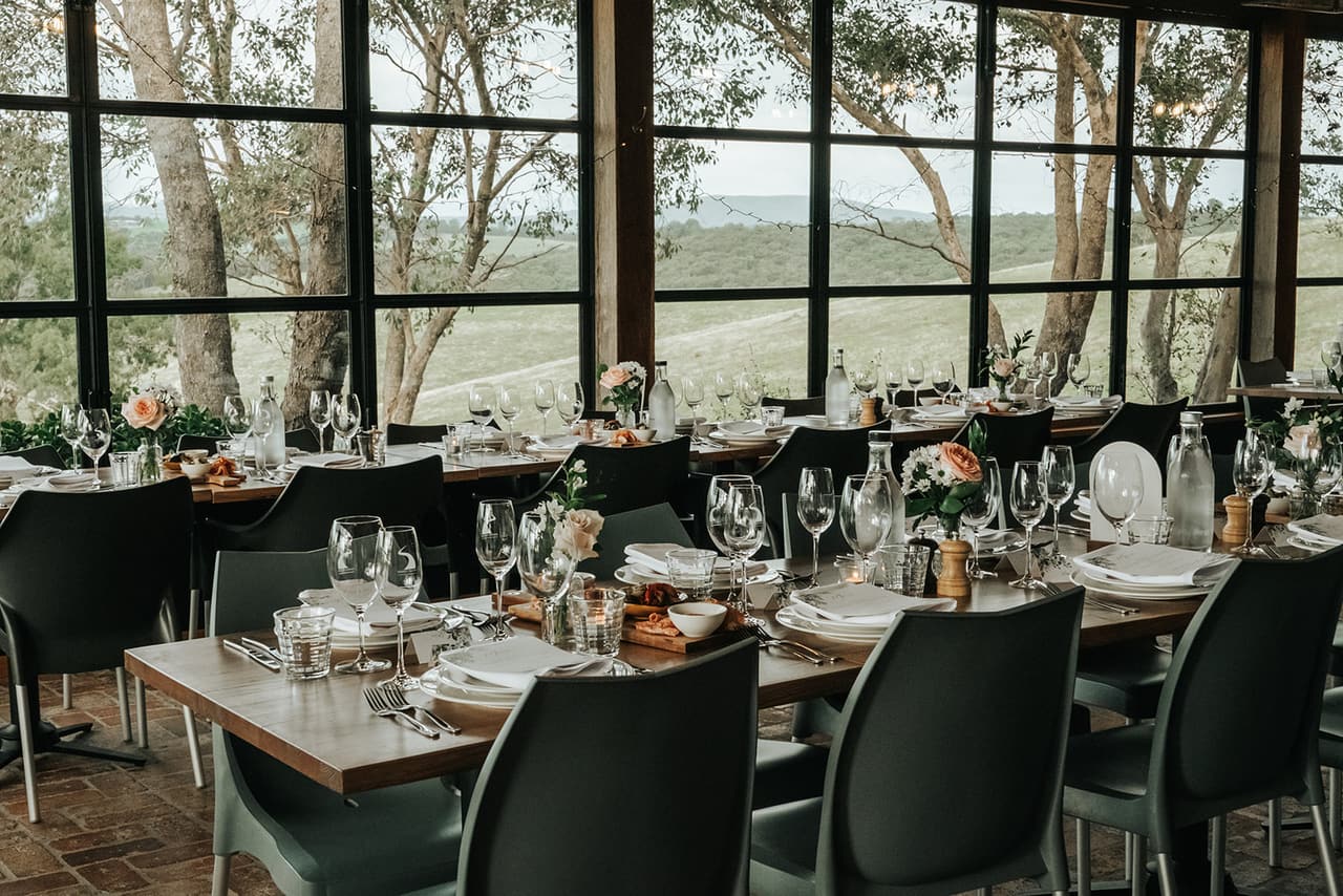 The reception room set for dinner, long timber tables and black chairs in front of tall black-framed windows over the hills