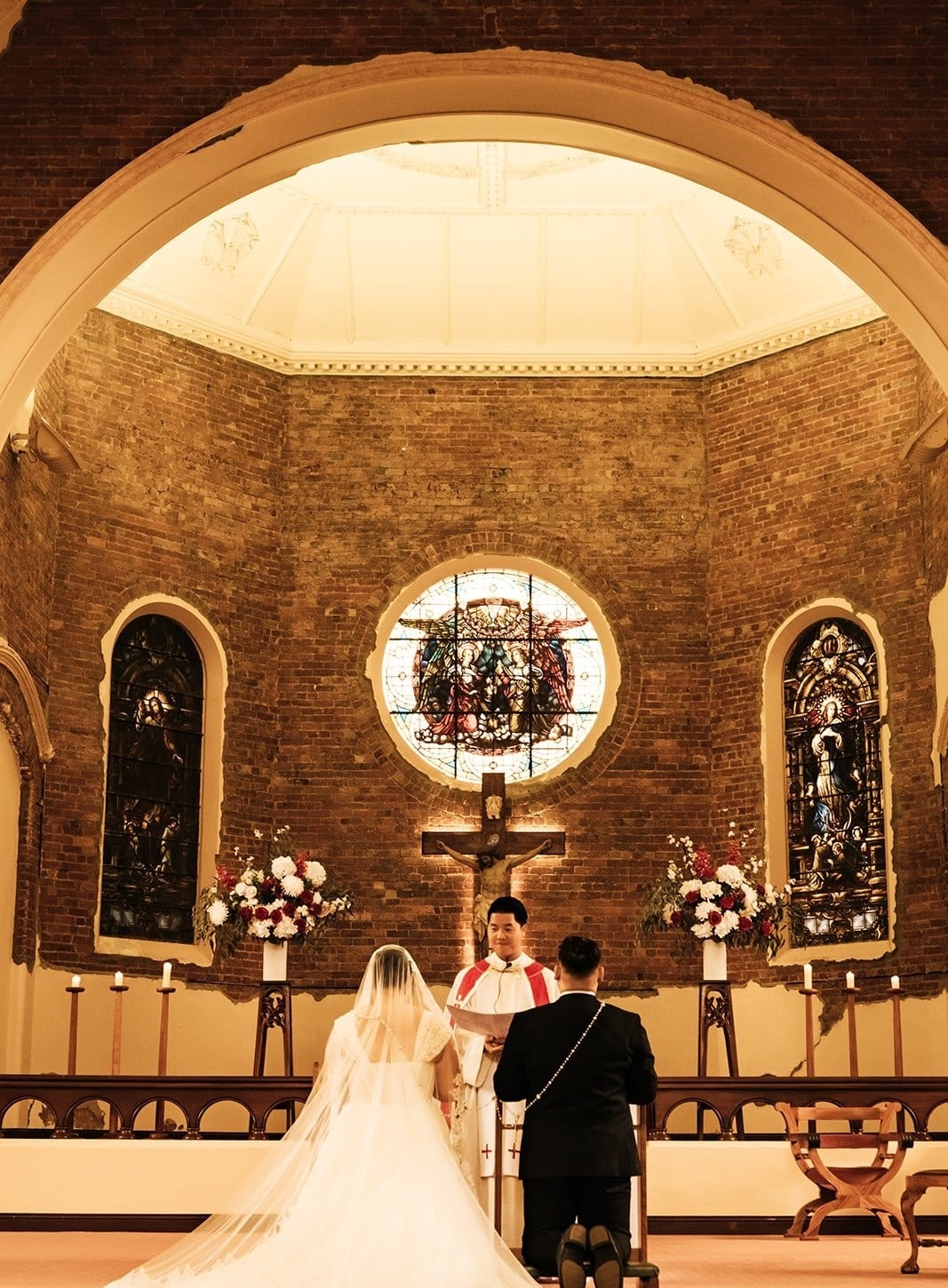 The couple kneeling before the altar under the round window, the veil spread on the floor