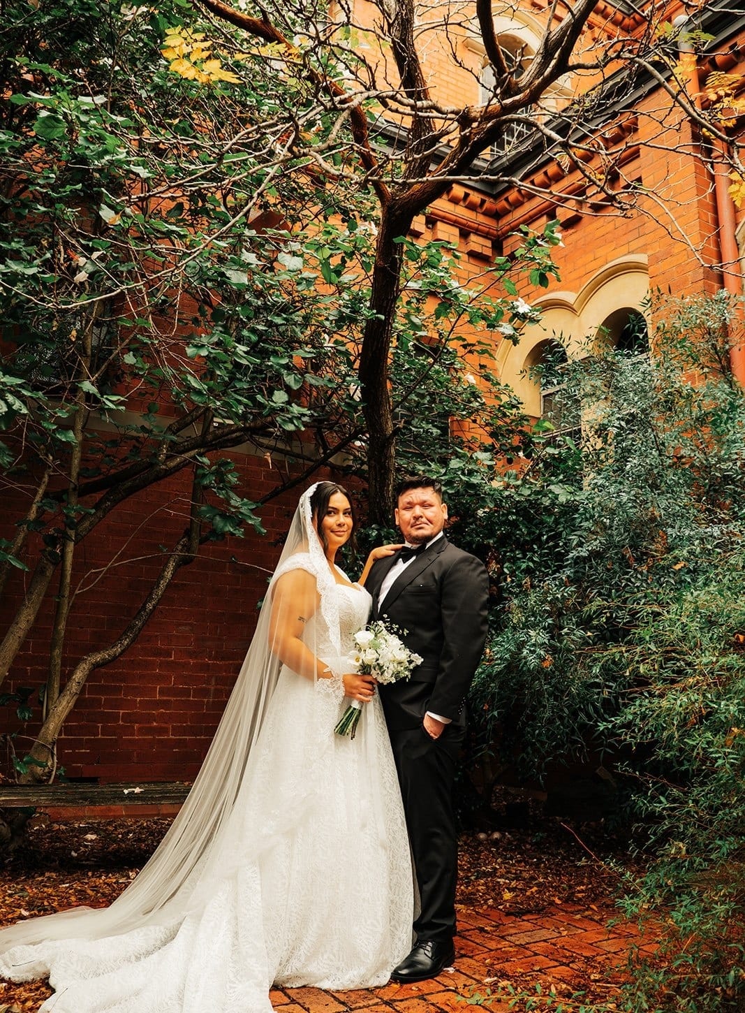 The couple under a tree in the garden beside the church, the brick walls behind them