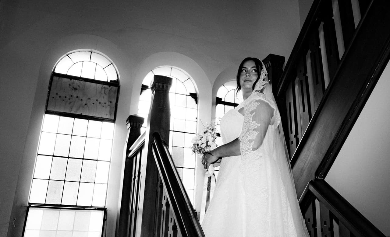 Black and white photo of the bride on the timber staircase beside tall arched windows
