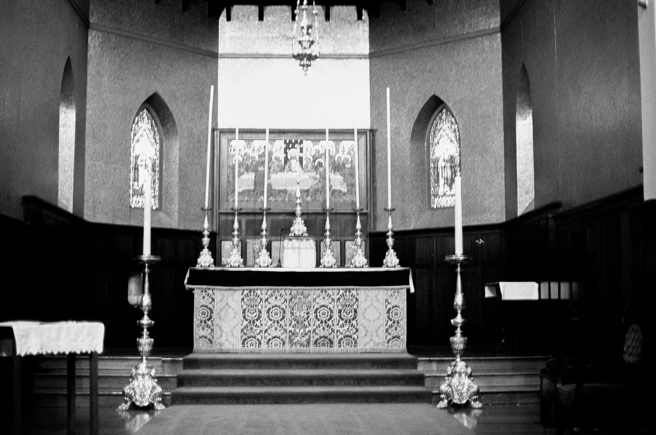 Black and white photo of the high altar with its tall candlesticks and the carved reredos behind, between two narrow stained glass windows, on 35mm film