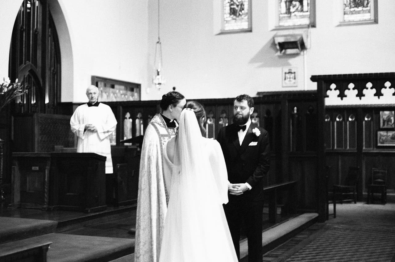 Black and white photo of Bianca and Aidan with the priest at the front of the church, a server in white standing by the dark timber choir stalls, on 35mm film