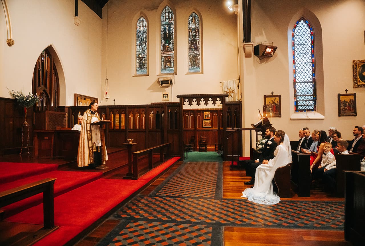 The front of the church from the side during the ceremony, the red carpeted sanctuary, the priest and Bianca and Aidan seated in the front pew