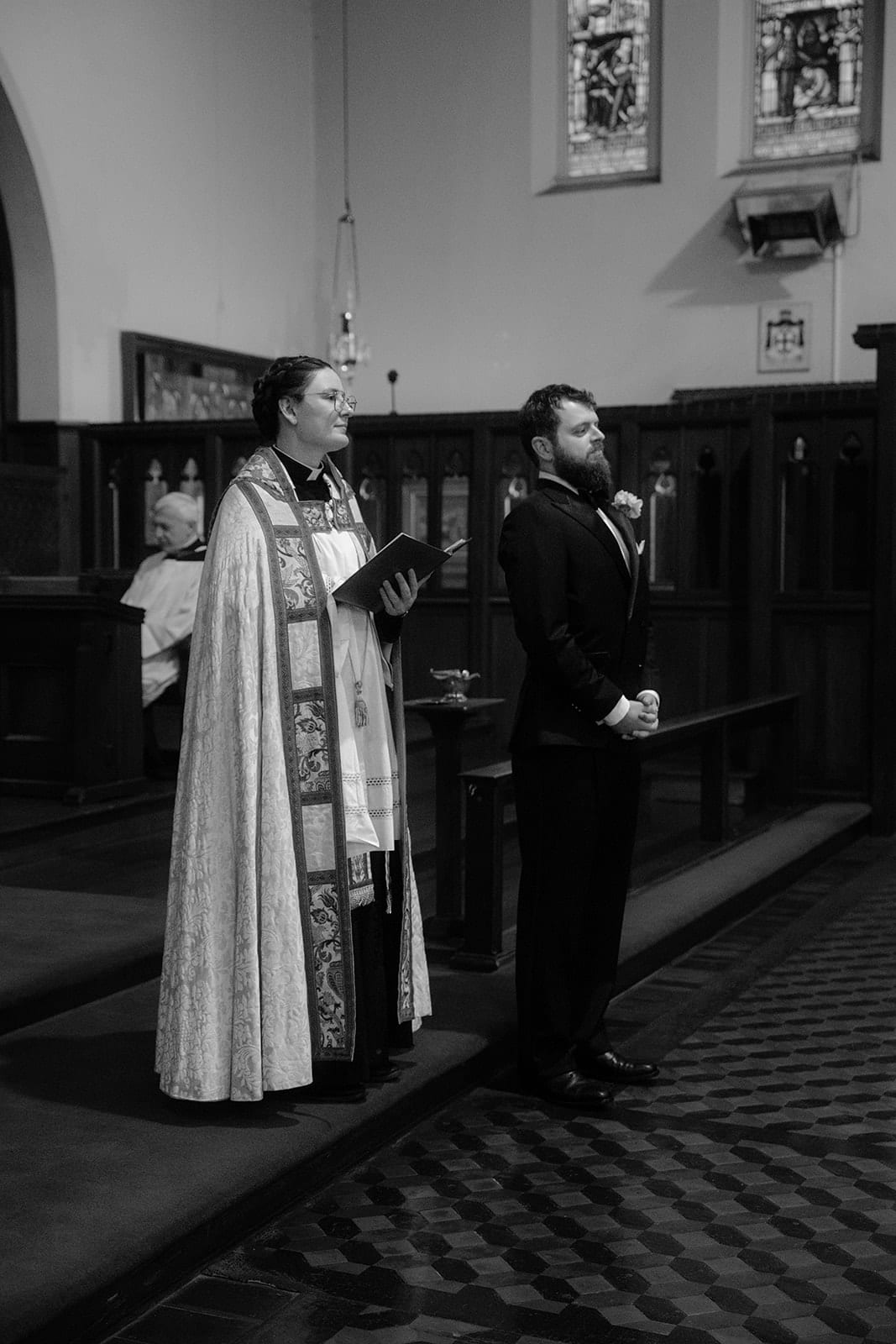 Black and white photo of the priest and Aidan waiting at the front of the church before the ceremony, dark timber choir stalls behind them