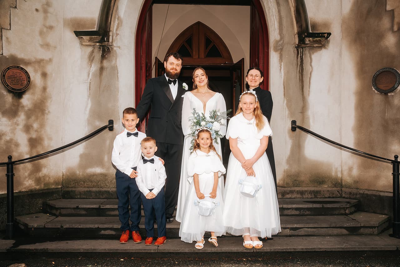 Bianca and Aidan with four children in white at the arched red timber door of the church, the stone steps and iron railings in front