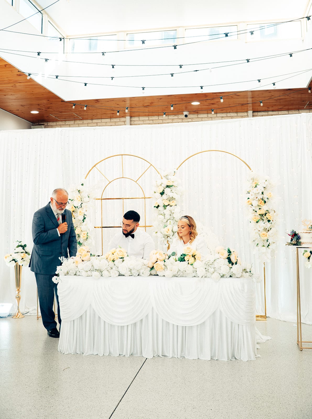 The hall from the back during the ceremony, an older man speaking in front of the head table under gold arches, festoon lights and skylights overhead