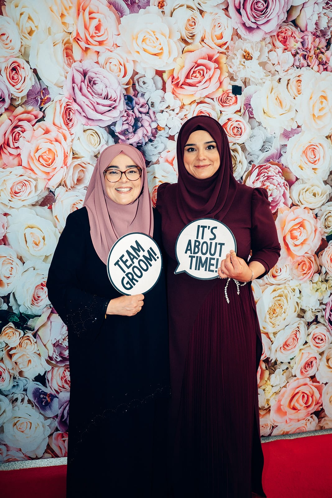 Two guests holding Team Groom and It’s About Time signs in front of a wall of pink, peach and white roses