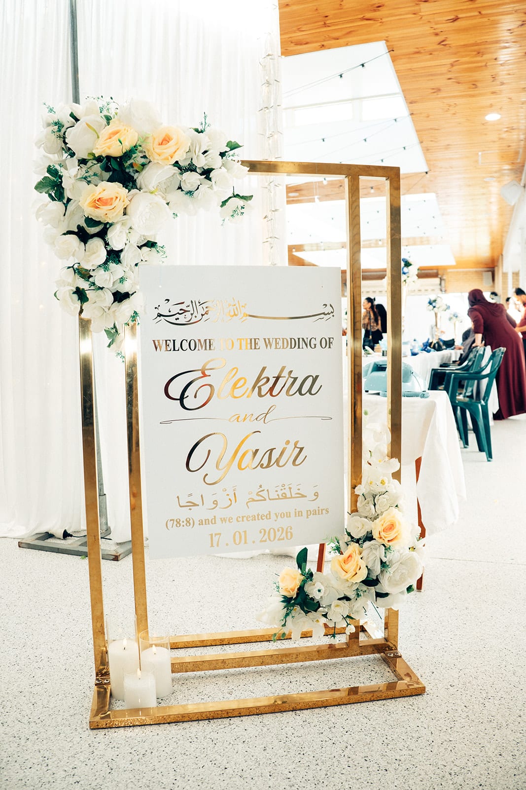 A white welcome sign for Elektra and Yasir with Arabic calligraphy and the date, on a gold frame with white and peach roses