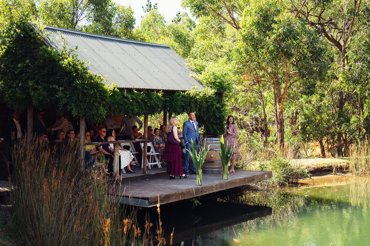 The Boatshed across the pond, a timber deck under a vine-covered roof, with guests seated for the ceremony