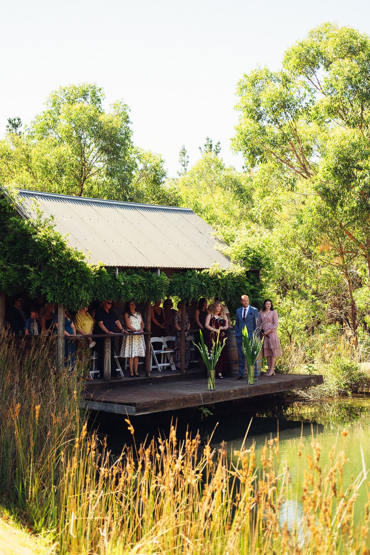 The Boatshed seen across the water through tall reeds, guests gathered on the deck before the ceremony