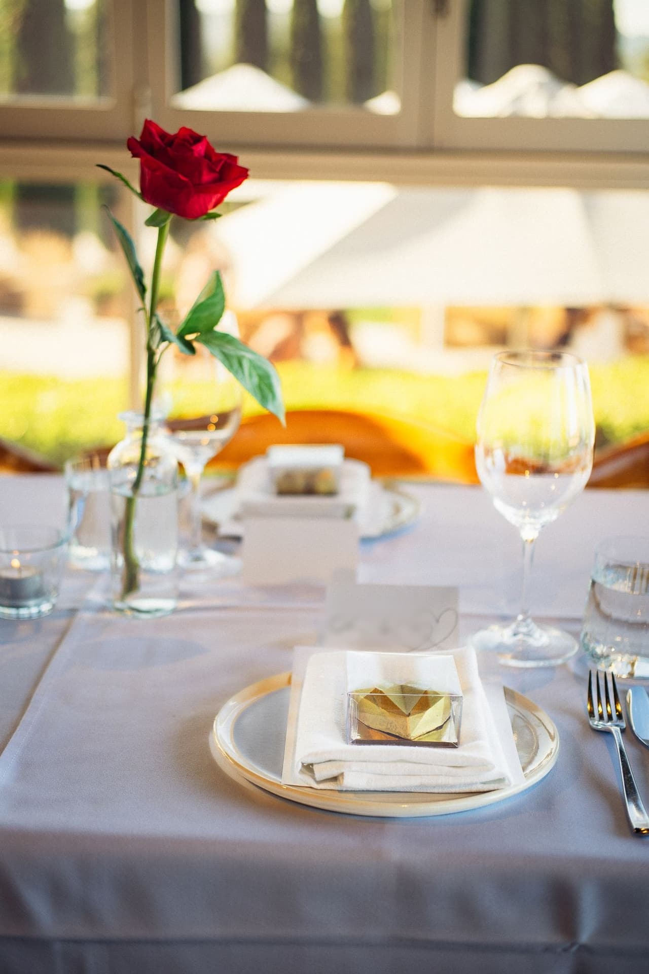 A single red rose in a bud vase on a white tablecloth beside a place setting and wine glasses