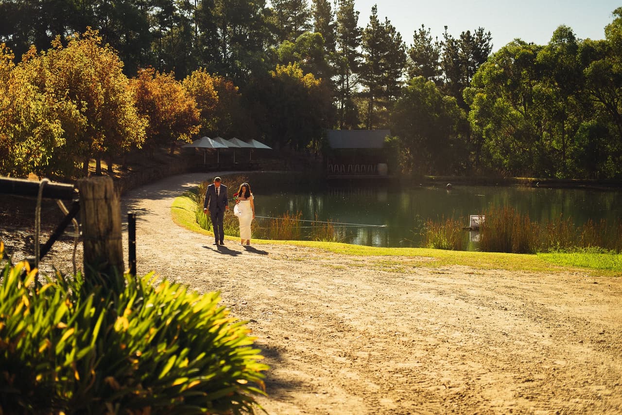 Kim and Ben walking along the dirt track beside the pond, the Boatshed and white umbrellas across the water