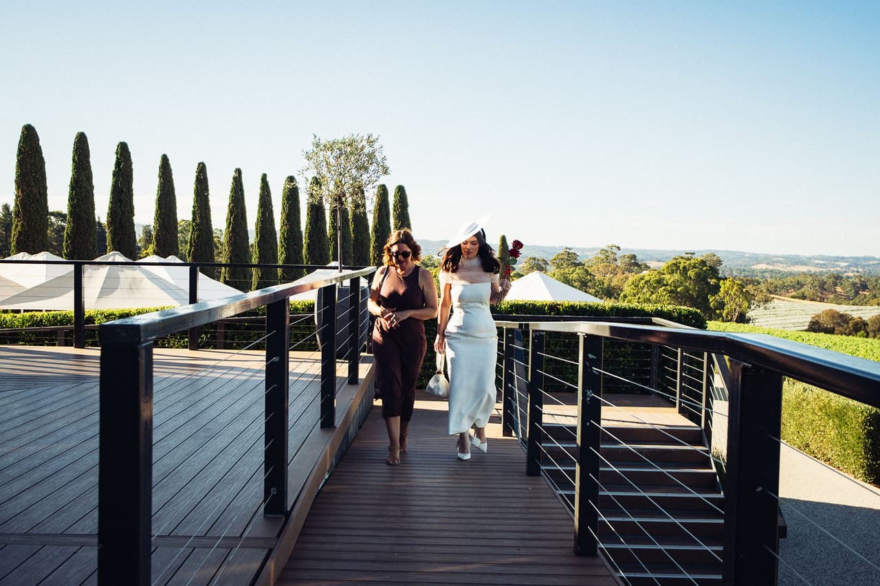 Kim walking with a guest along the boardwalk towards the restaurant, a row of cypresses and the valley behind