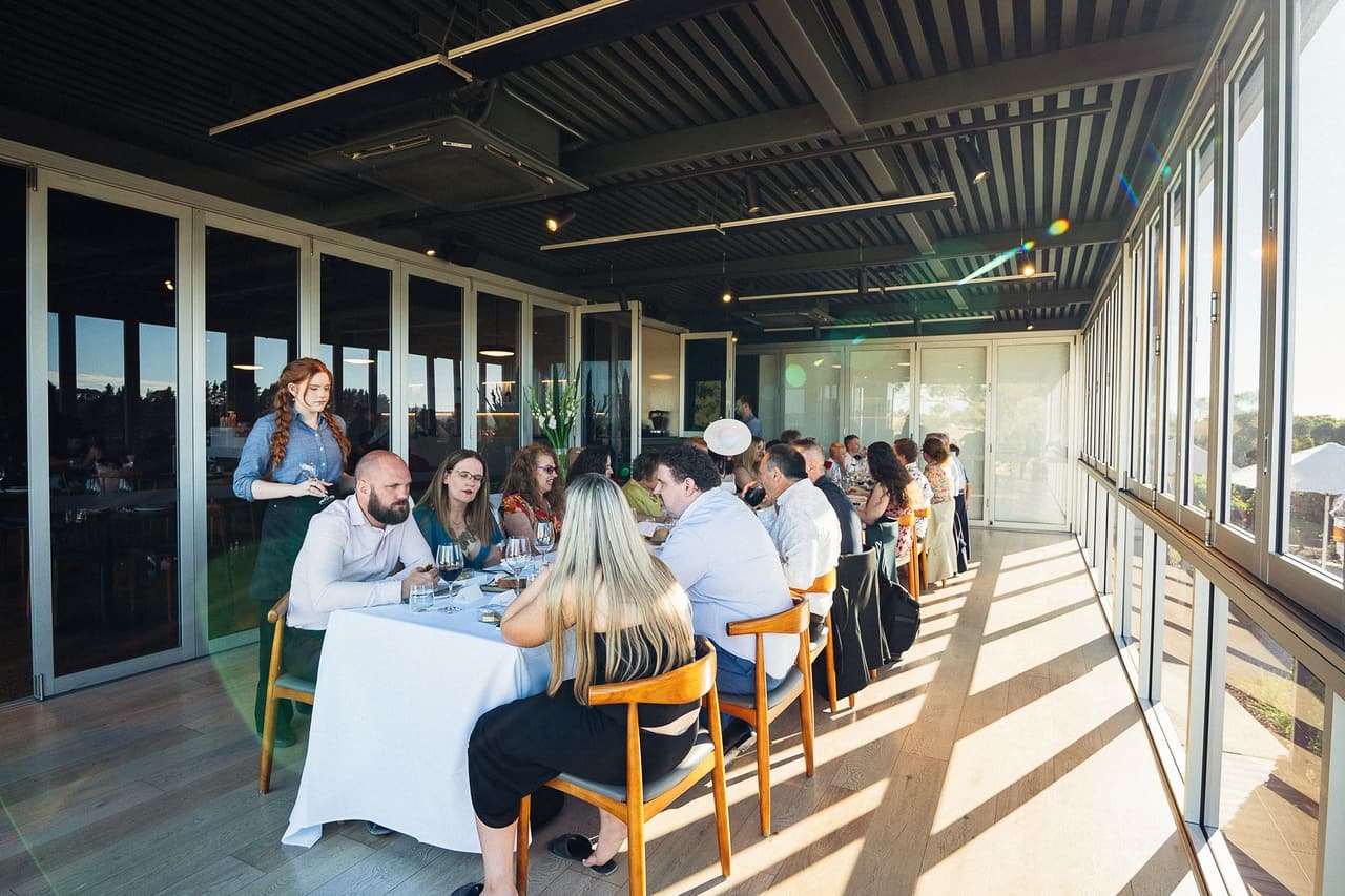 The restaurant from the end of the room, guests at long white tables and the sun falling through the glass walls