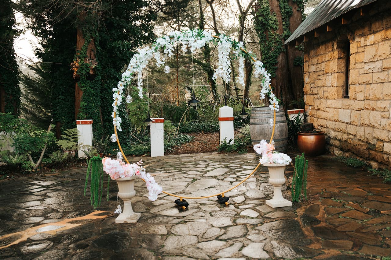A pink and white floral arch on the flagstones beside the old stone cottage, ready for the vows