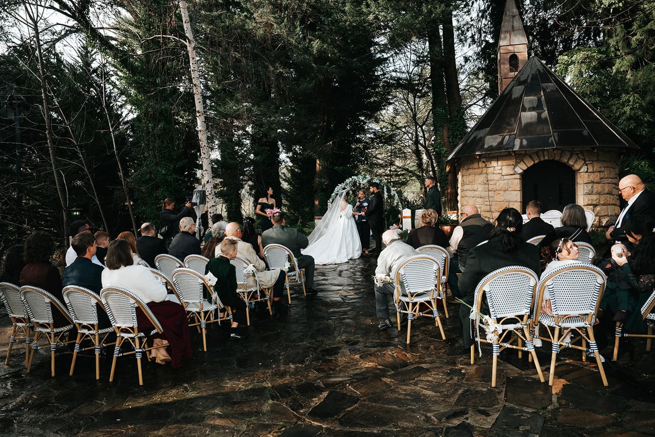 Guests seated under the trees in the drizzle as the ceremony begins by the garden chapel