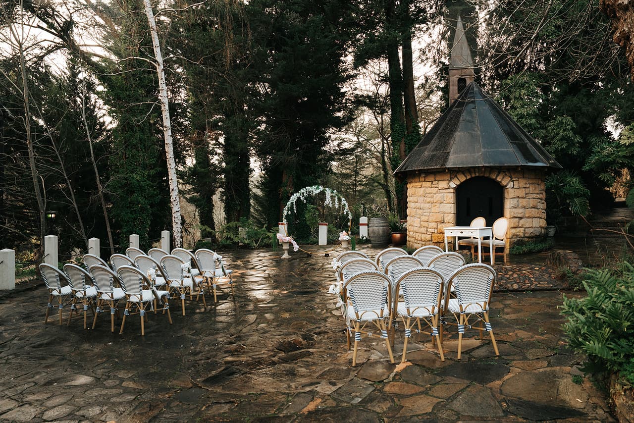 The ceremony set up beside the little stone chapel, white cane chairs on wet flagstones under tall trees