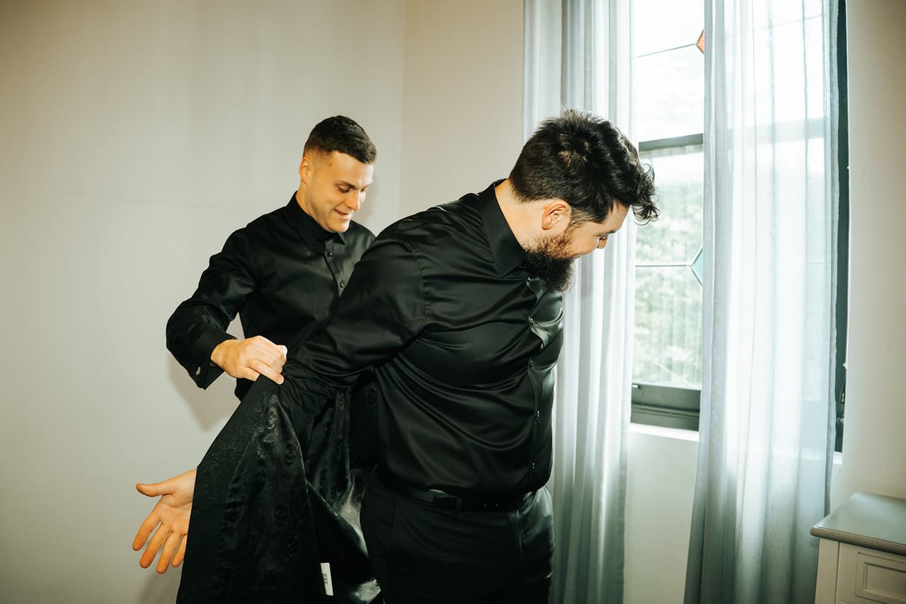 A groomsman helping Jordan with his shirt by the tall windows of the groomsmen’s room