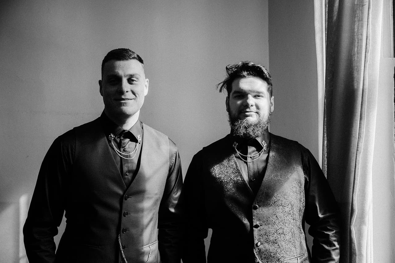 Black and white portrait of Jordan and a groomsman in their dark suits beside the window