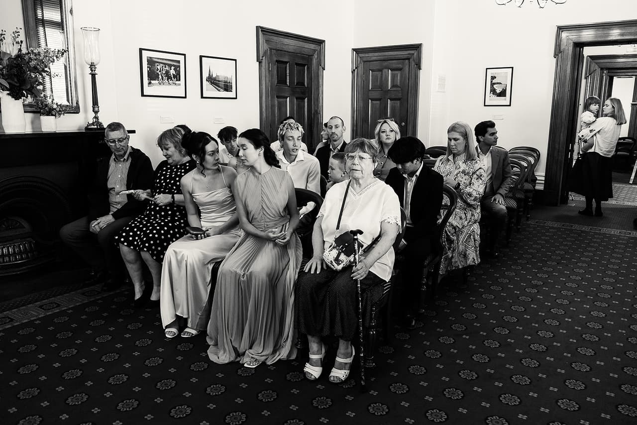 Black and white photo of guests waiting in their seats along the side wall of the Margaret Craig Room before Cecile and Stefan’s ceremony