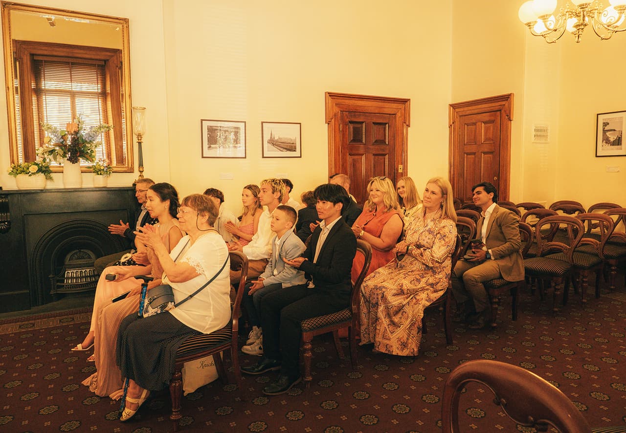 Guests seated in rows of timber chairs in the Margaret Craig Room, a dark fireplace with flowers on the left and Cecile and Stefan at the registry desk at the far end