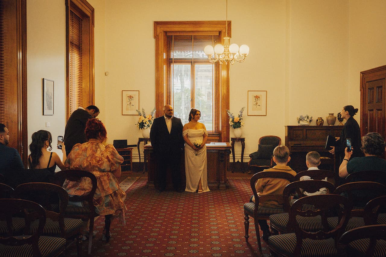 Martina and Diego’s ceremony seen from behind a few guests, the couple at the registry desk under the window and the celebrant reading beside them