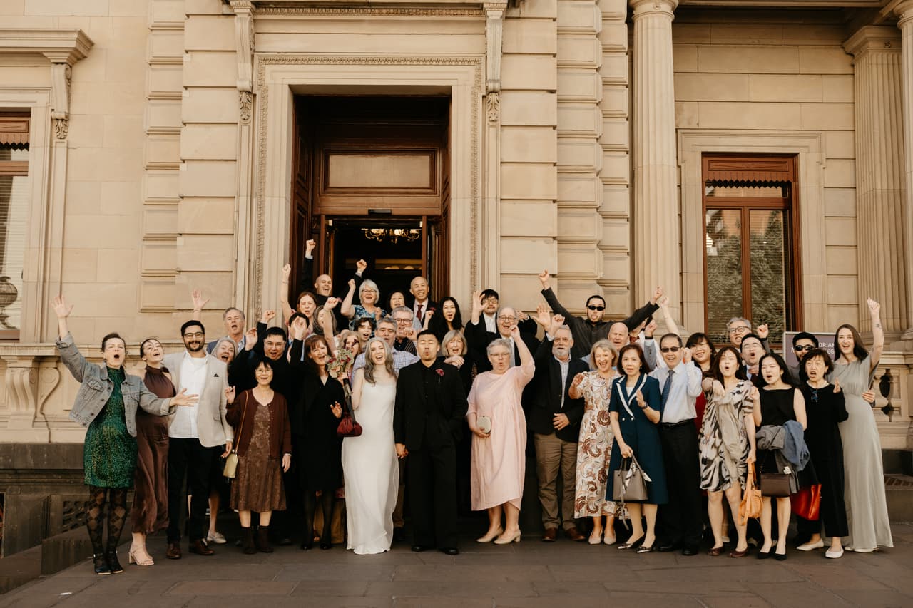A big group of family and friends cheering with the bride and groom outside the registry entrance after the ceremony, the sandstone facade, columns and tall timber doors of the Old Treasury Building behind them