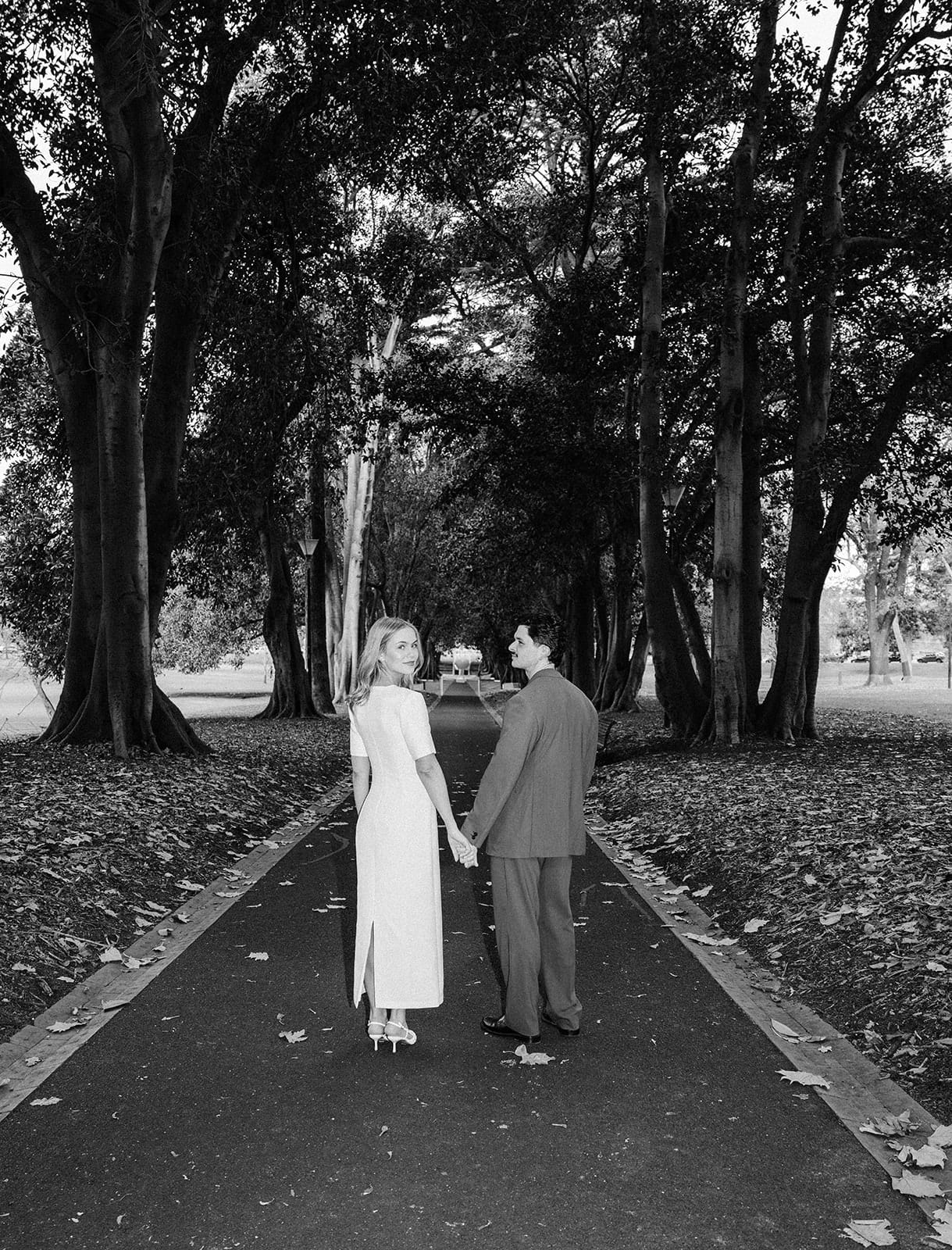 Black and white photo of Karina and Chris hand in hand on a path under an avenue of old trees in Treasury Gardens, Karina looking back