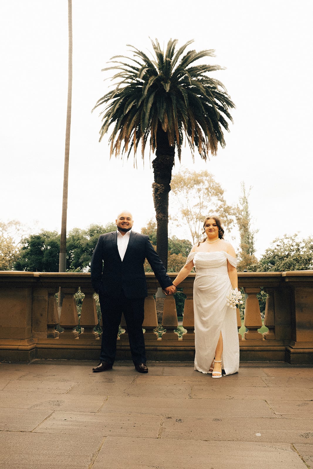 Martina and Diego holding hands on the sandstone balcony of the Old Treasury Building, a tall palm and the gardens beyond the balustrade
