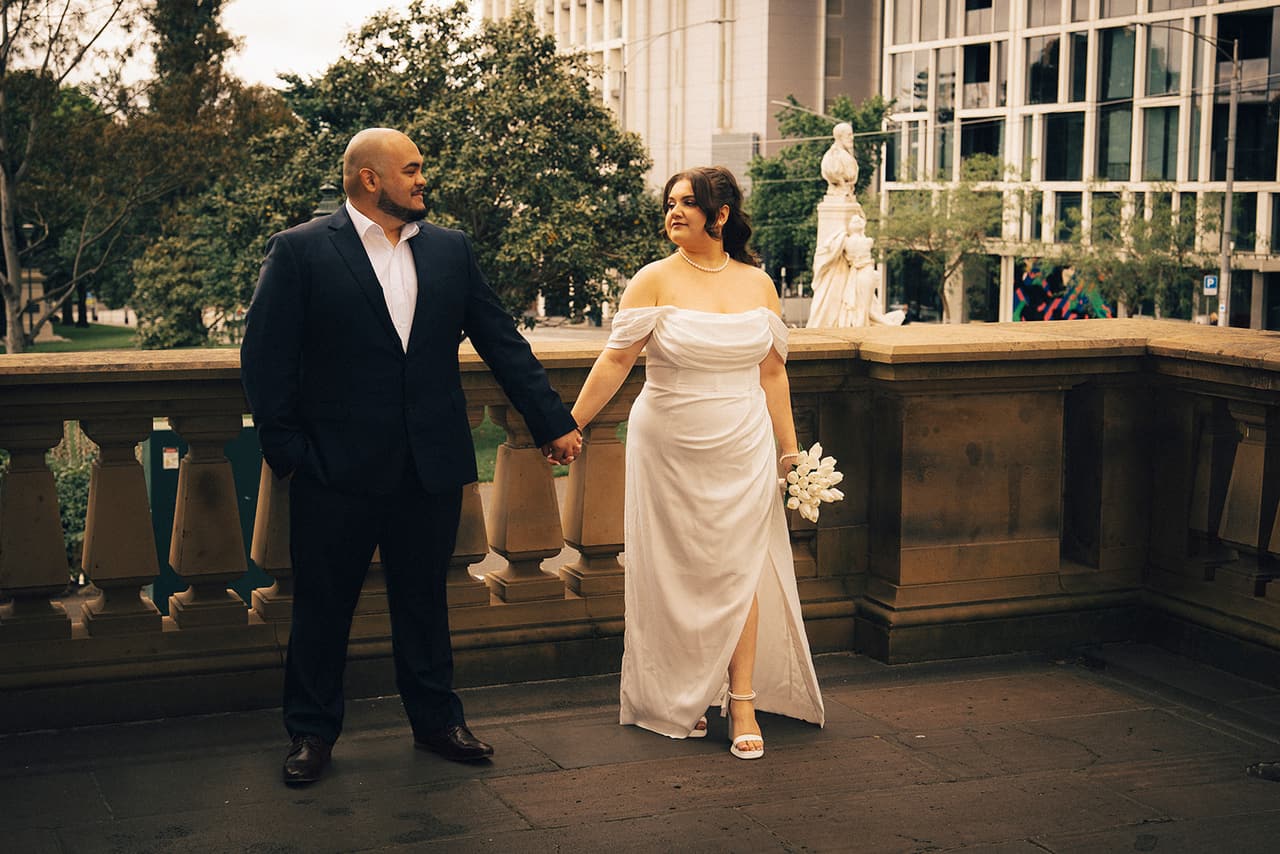 Martina and Diego holding hands at the end of the balcony, a white statue on the balustrade and office towers beyond