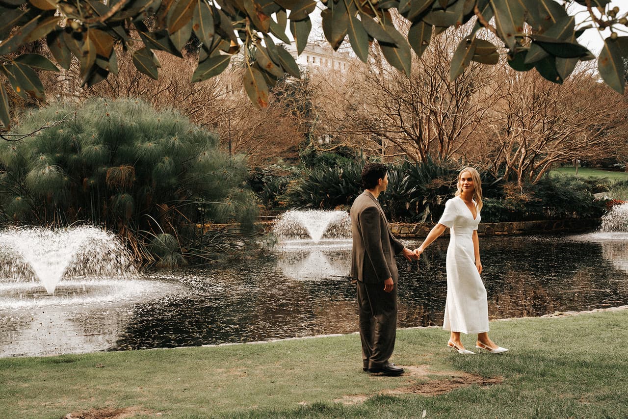 Karina and Chris holding hands beside the pond and its fountains, framed by overhanging branches