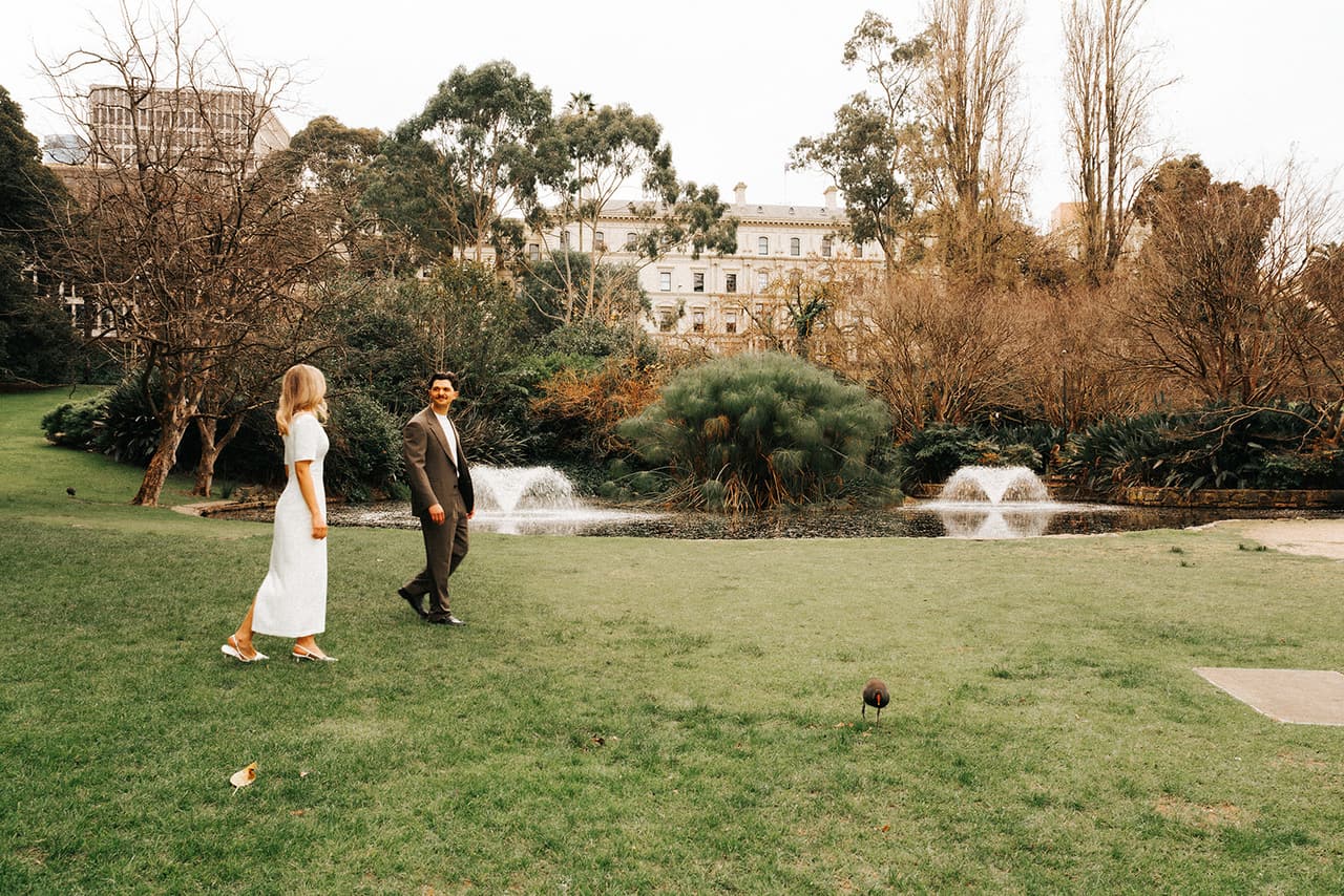 Karina walking towards Chris on the lawn by the pond in Treasury Gardens, two fountains playing and bare winter trees behind