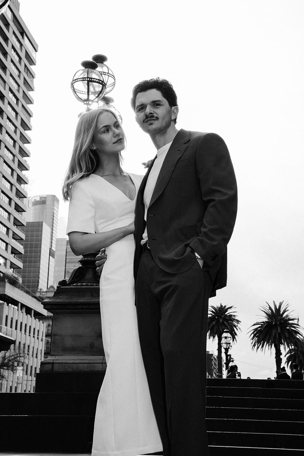 Black and white photo of Karina and Chris at the top of the stone steps beside an old lamp standard, city towers and palm trees behind them