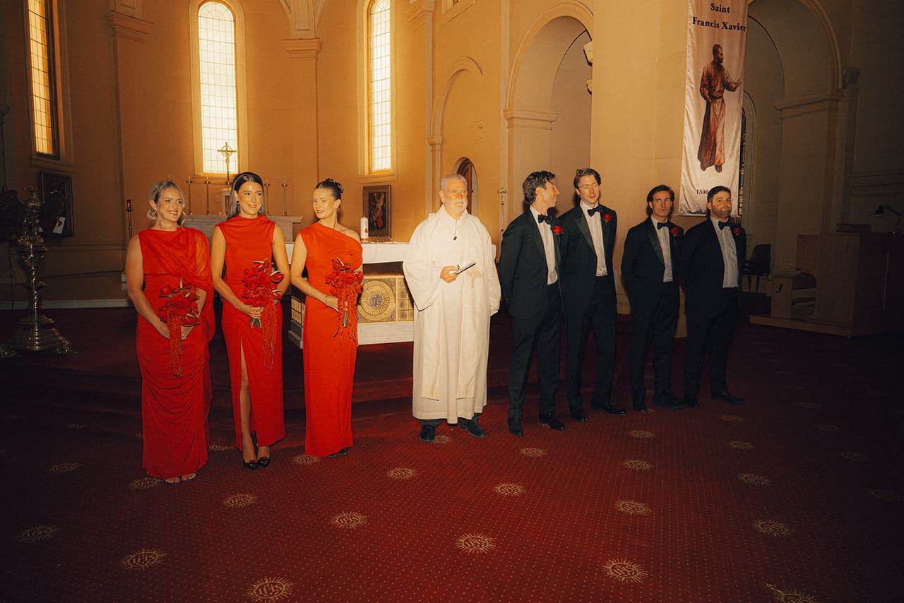 Bridesmaids in red and groomsmen in black tie waiting either side of the priest at the front of the chapel