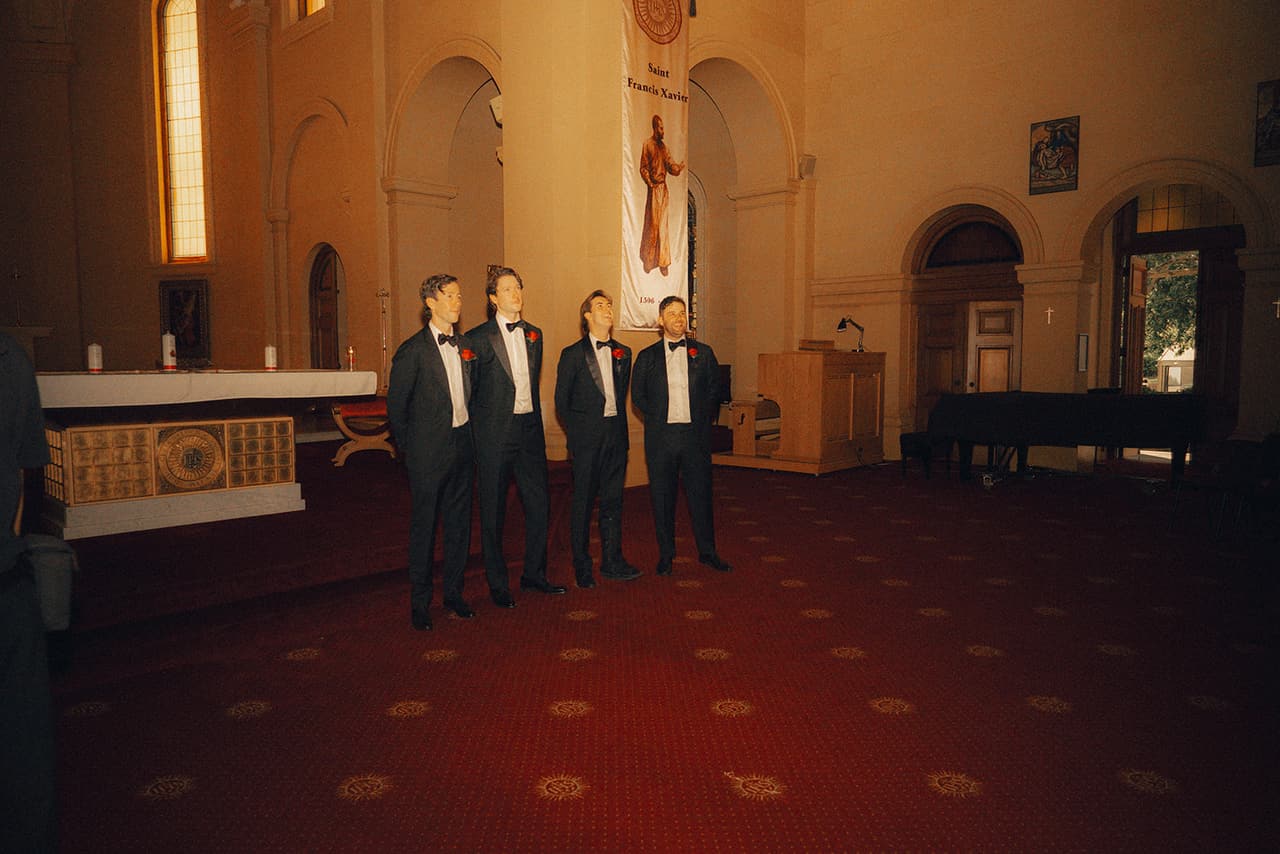 Four men in black tie with red rose buttonholes waiting on the red carpet at the front of the chapel beside the altar