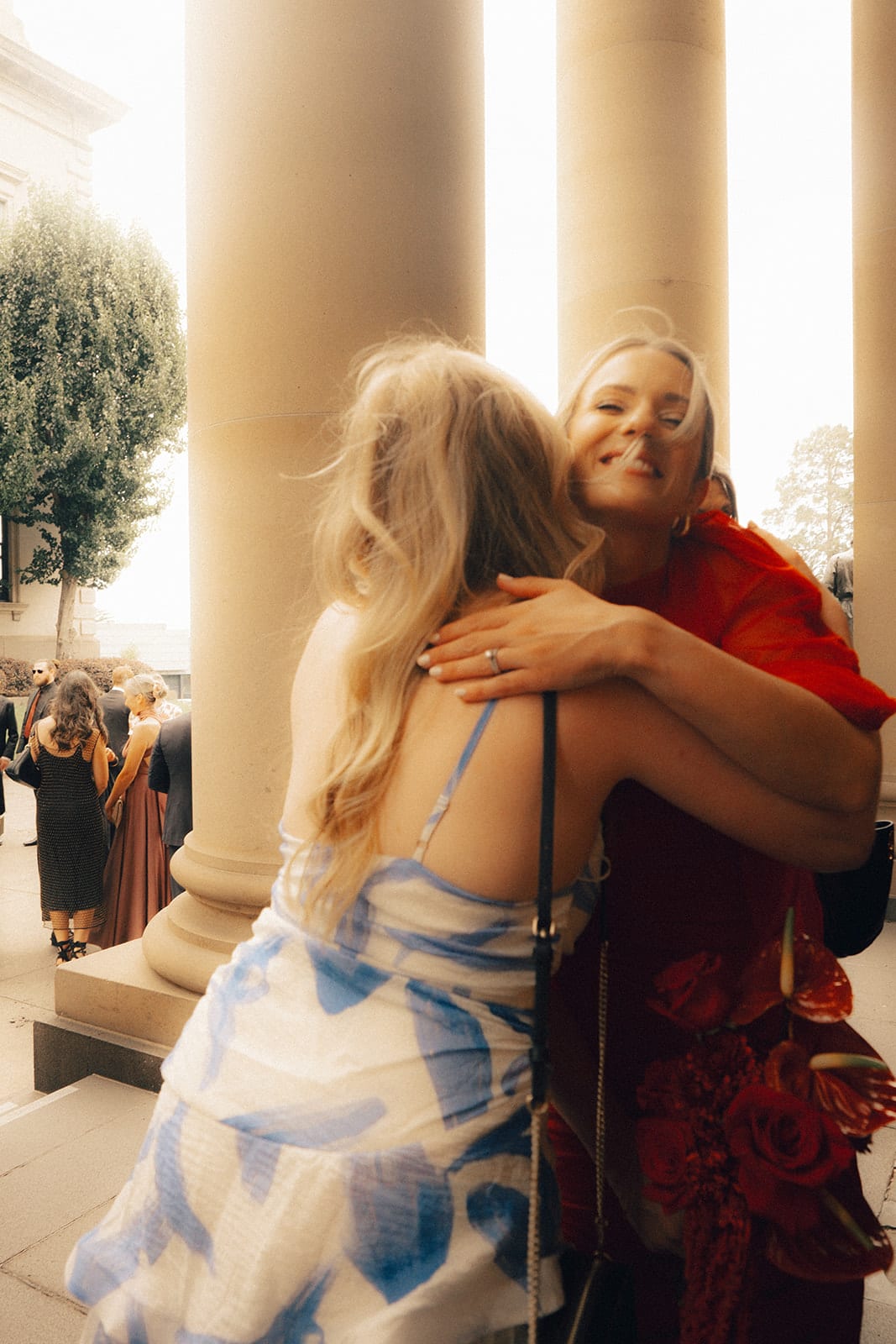 A bridesmaid in red hugging a guest between the sandstone columns of the chapel portico