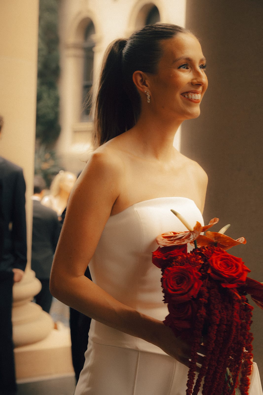 Kira smiling beside a sandstone column of the chapel portico, her red bouquet in her hands