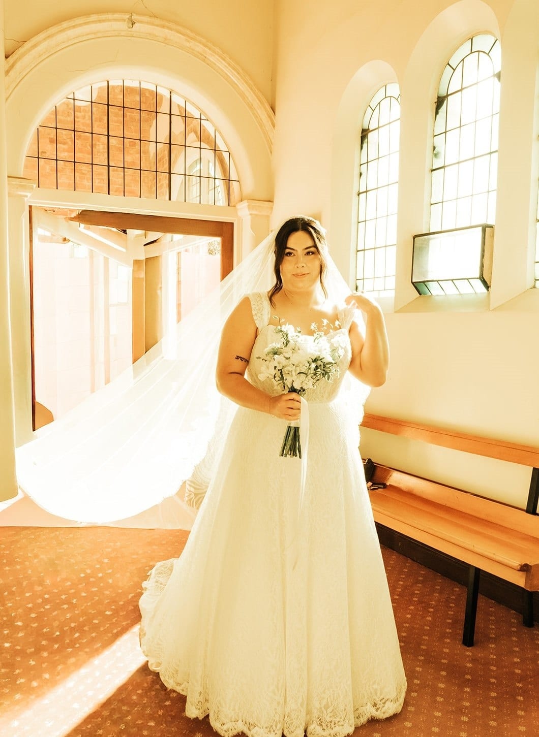 Ashley in her lace gown and long veil holding white flowers at the back of the church