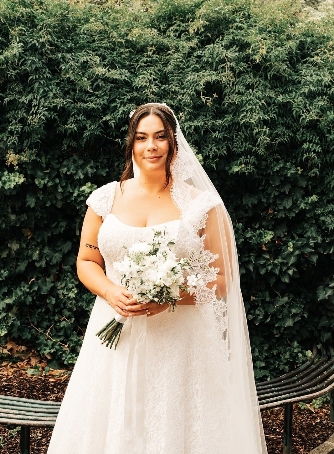Ashley in her veil holding white flowers in front of a green hedge