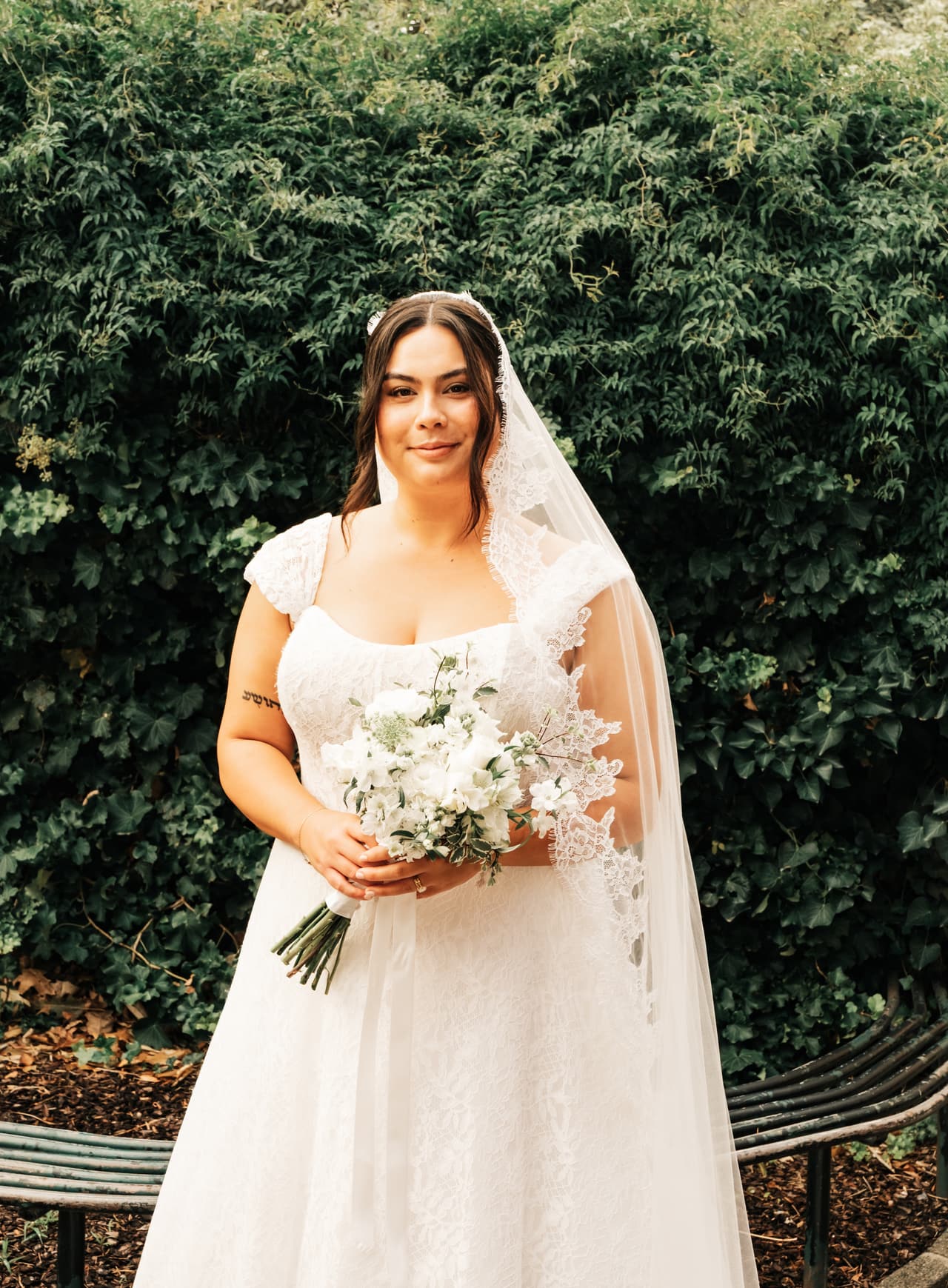 Ashley in her veil holding white flowers in front of a green hedge