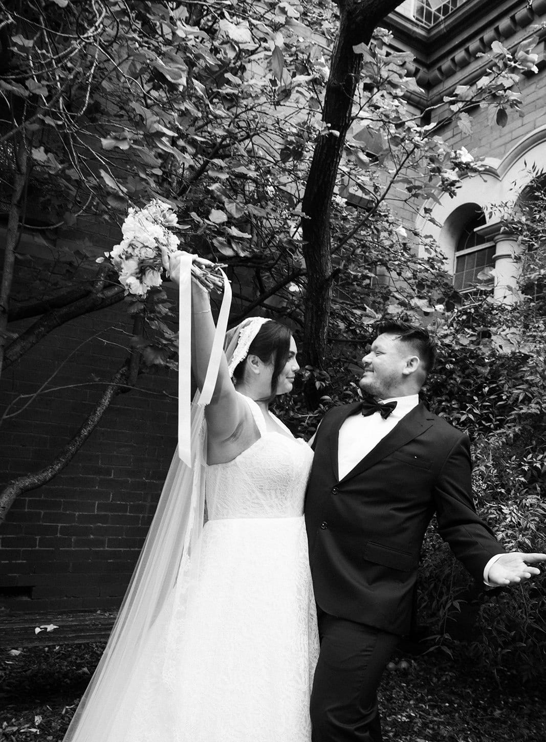 Black and white photo of Ashley raising her bouquet as Bryan laughs beside her
