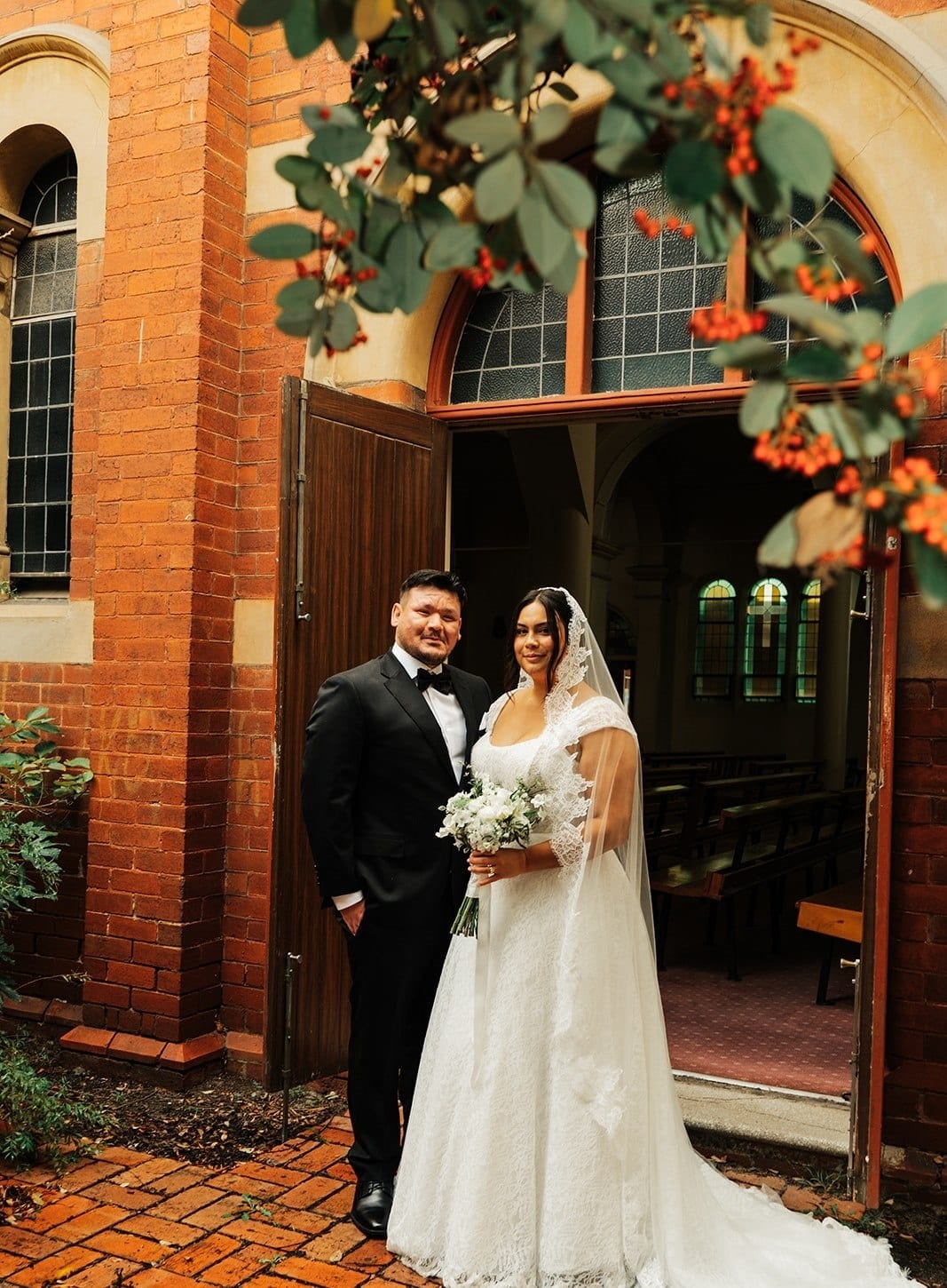 Ashley and Bryan standing at the open timber doors of the red brick church