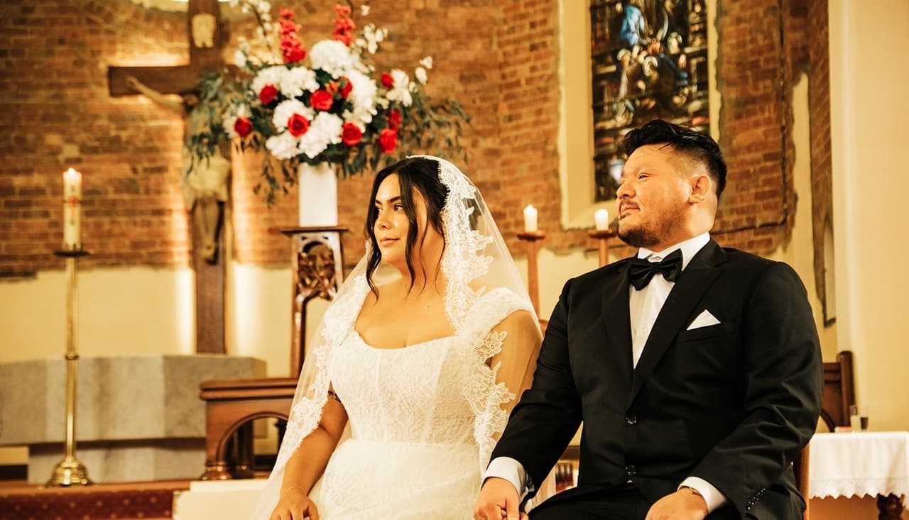 Ashley and Bryan seated side by side in the sanctuary under red and white flowers